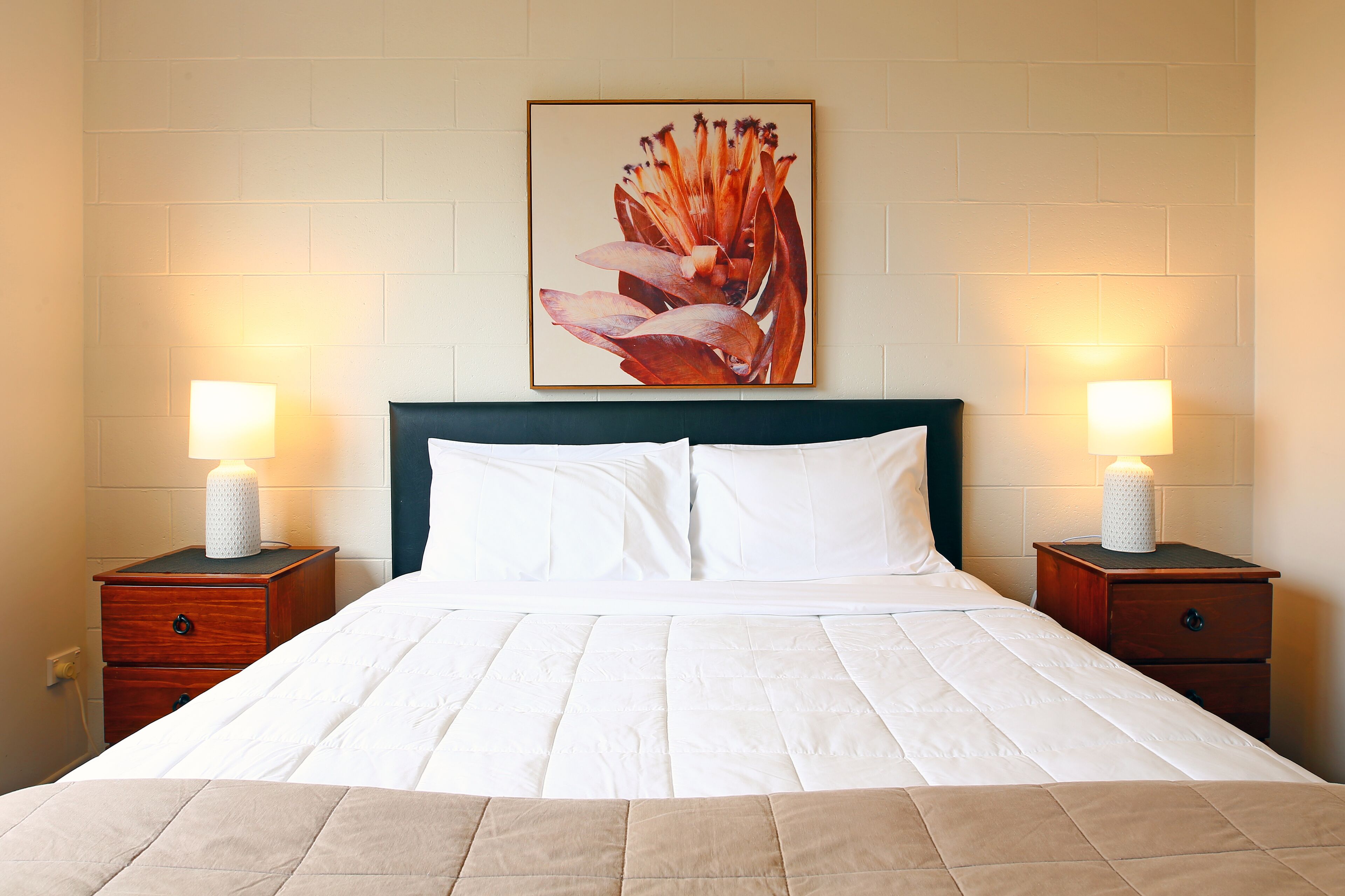 A neatly made bed with white bedding, flanked by two bedside tables with lamps, in a room with a white brick wall and a framed floral artwork above the bed.