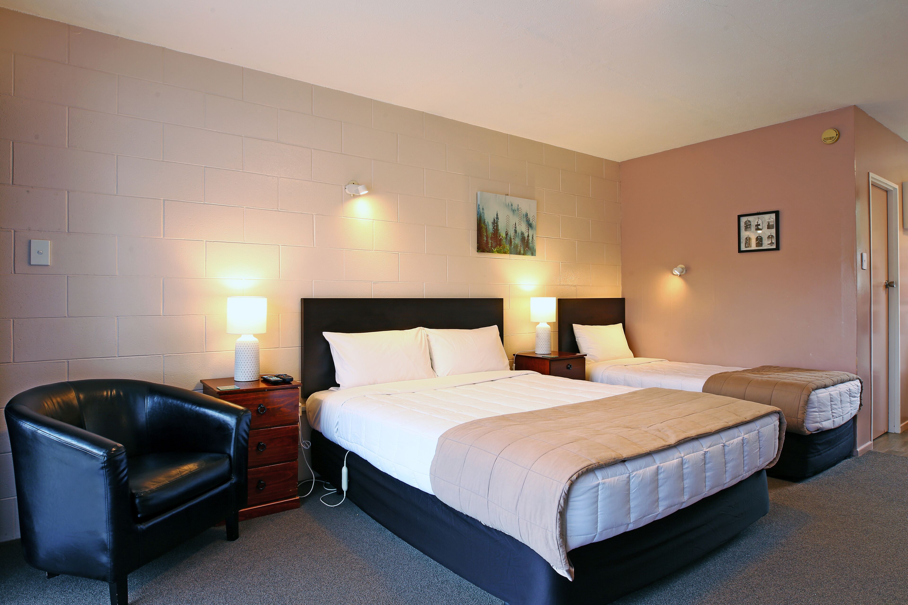 Une chambre d’hôtel avec deux lits, un fauteuil en cuir noir et un petit bureau avec une lampe.