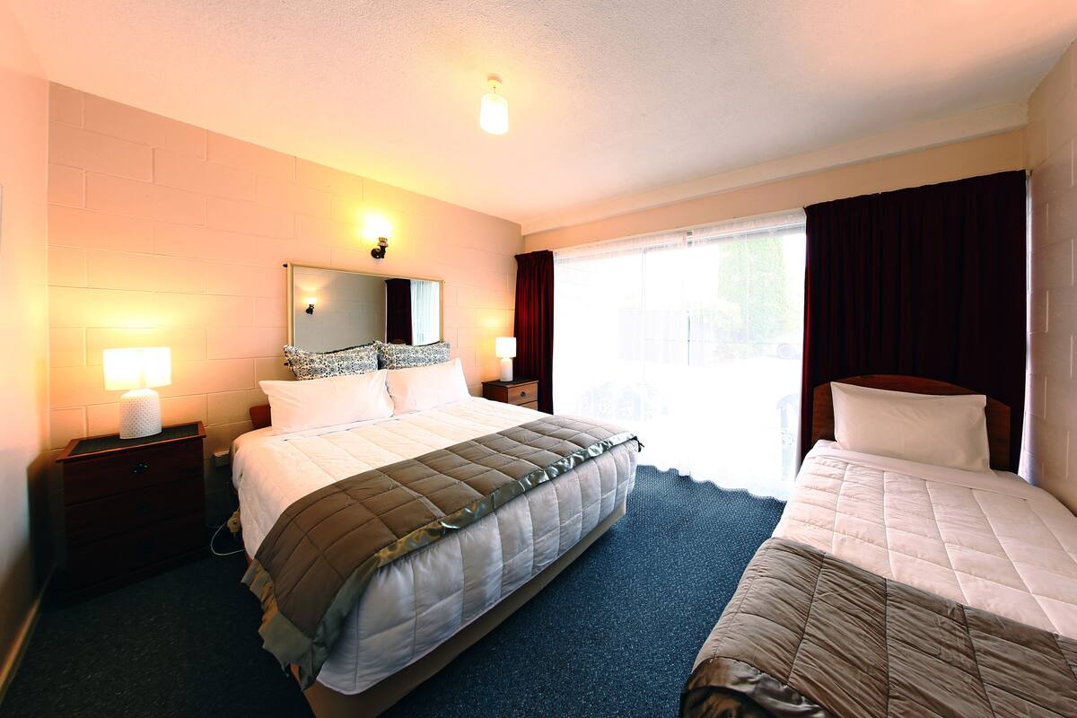 A hotel room with a large bed, a smaller bed, a nightstand with a lamp, a mirror, and a window with curtains.