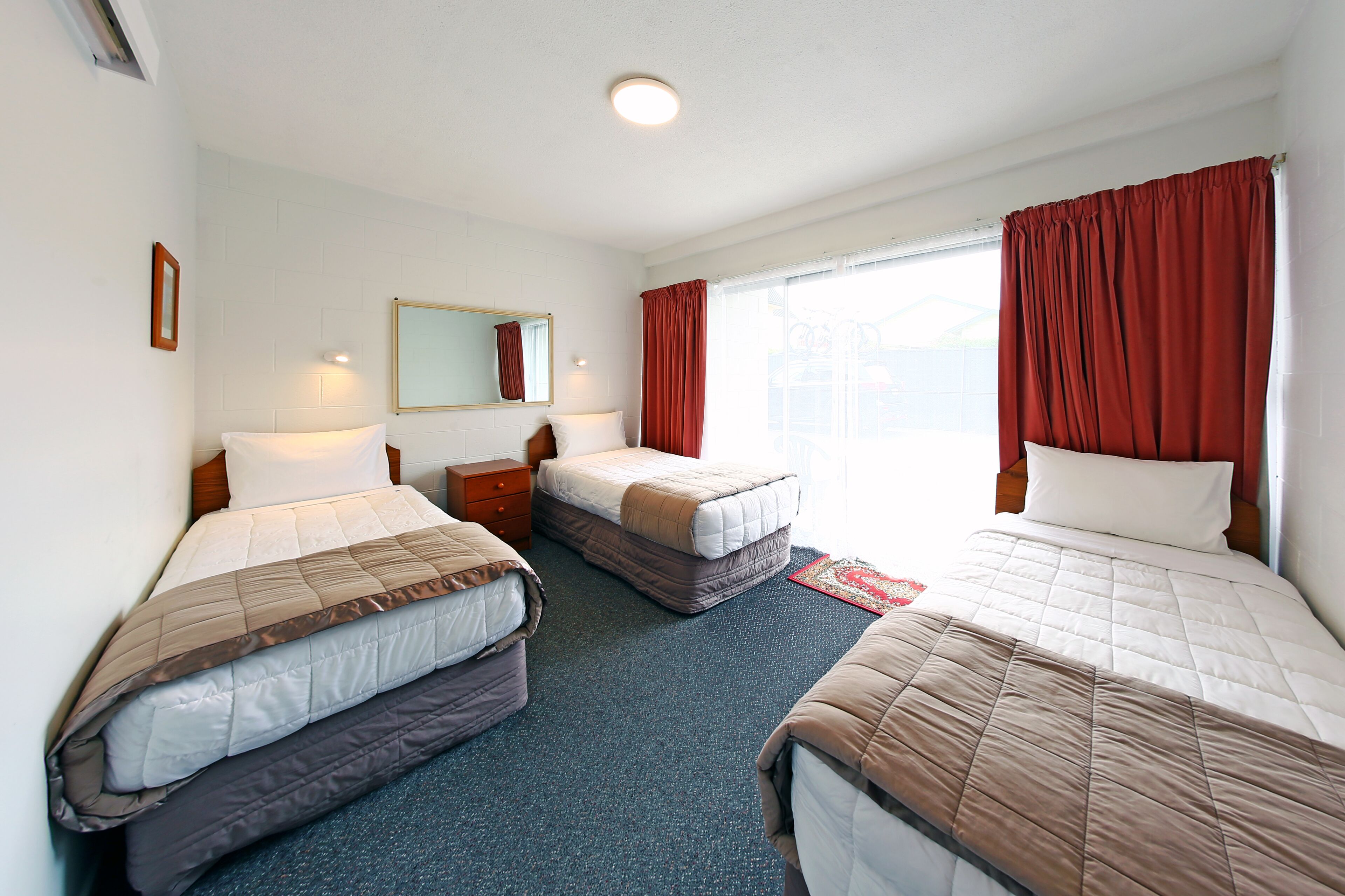 A hotel room with three beds, a small bedside table, a mirror, and a window with red curtains.