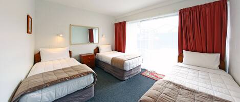 A hotel room with three beds, a small bedside table, a mirror, and a window with red curtains.