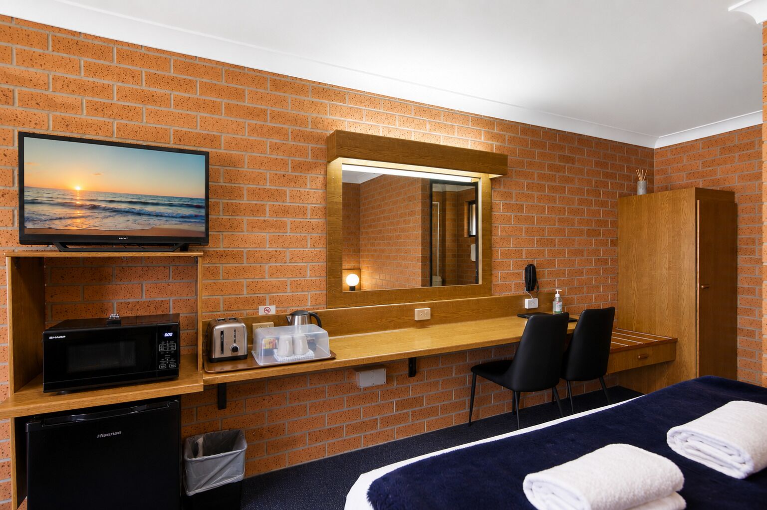 Habitación de hotel con pared de ladrillo, televisor, microondas, lavamanos, espejo, cama con toallas y escritorio con silla.