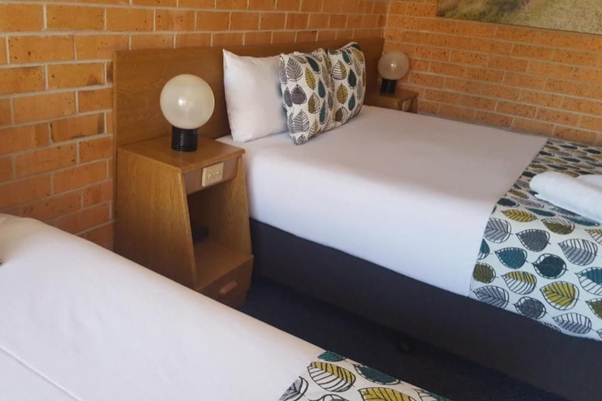 Habitación de hotel con dos camas, un muro de ladrillo y una mesita de noche con lámpara.
