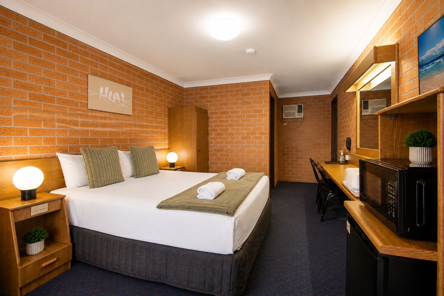 A hotel room with a brick wall, a bed with pillows, a bedside table with a lamp, a desk with a chair, and a microwave.