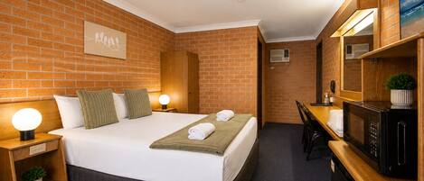 A hotel room with a brick wall, a bed with pillows, a bedside table with a lamp, a desk with a chair, and a microwave.