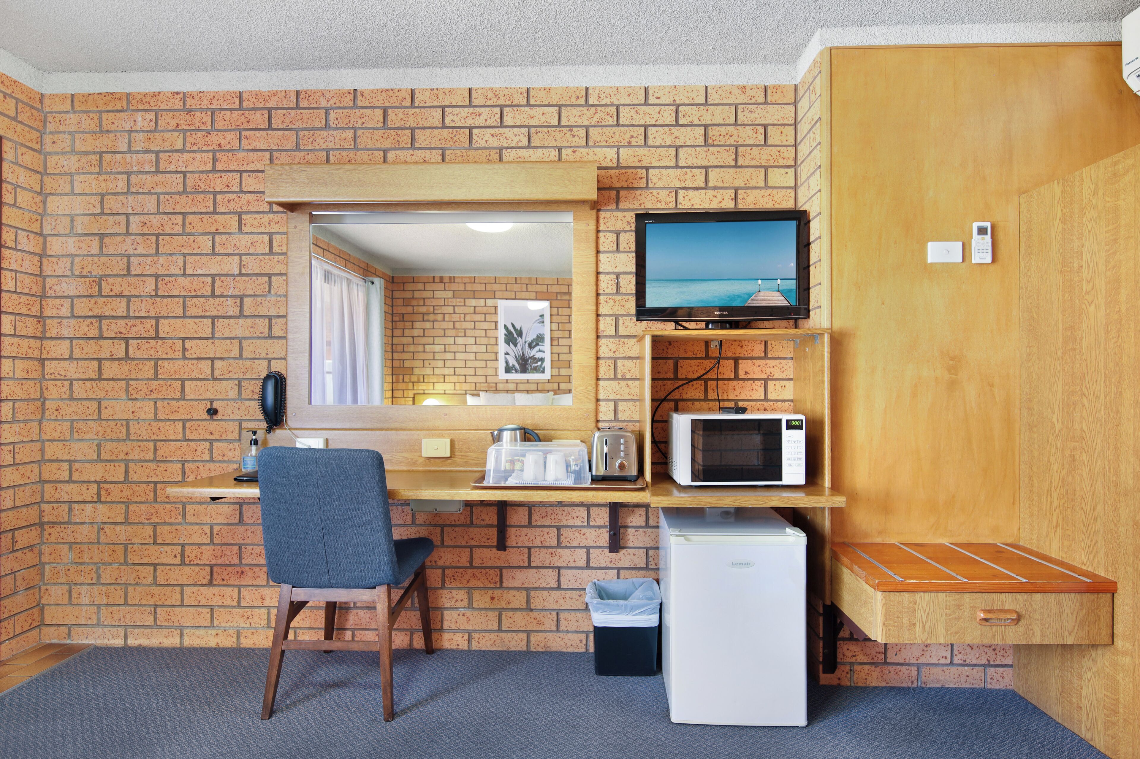 Una habitación funcional con pared de ladrillo, un escritorio de madera, una silla, televisor, microondas, refrigerador y un bote de basura.