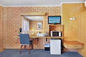 A compact room with a brick wall, a wooden desk, a chair, a television, a microwave, a refrigerator, and a trash bin.