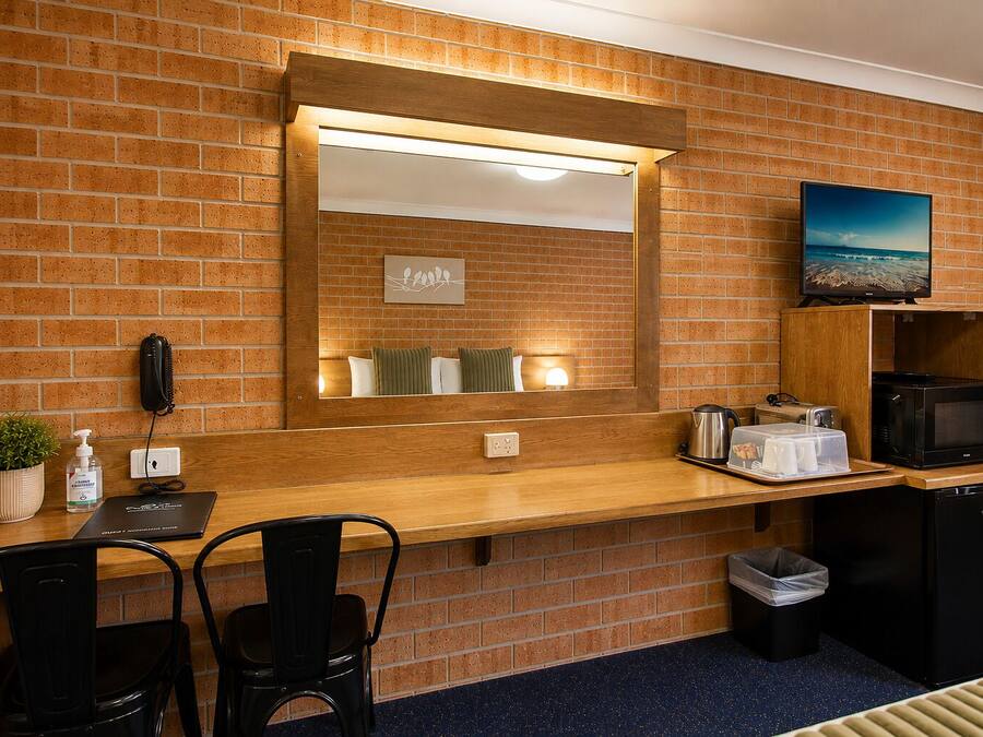 Quarto de hotel com escrivaninha de madeira, espelho, micro-ondas, lixeira e uma pequena planta.
