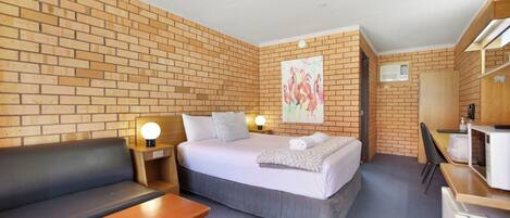A hotel room with a brick wall, a bed with white bedding, a wooden desk, a black leather sofa, and a painting of flamingos.