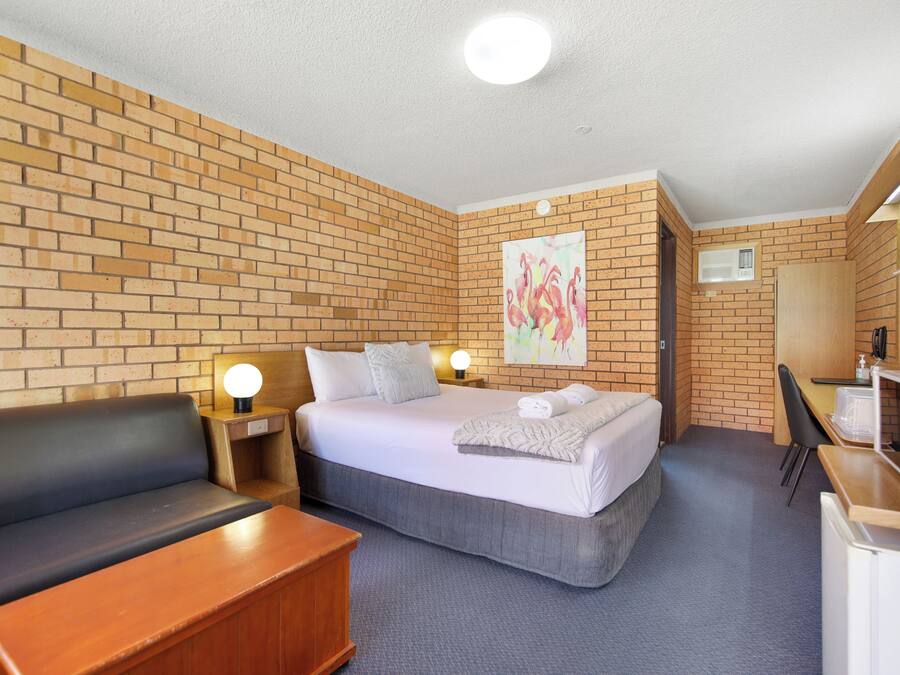 A hotel room with a brick wall, a bed with white bedding, a wooden desk, a black leather sofa, and a painting of flamingos.