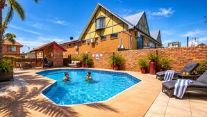 A swimming pool with two people, surrounded by lounge chairs and potted plants, with a brick building and a clear sky in the background. - Royal Palms Motor Inn (Coffs Harbour)