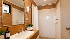 Un baño con tocador de madera, un lavabo blanco, un espejo, un secador de pelo montado en la pared y una zona de ducha con piso de baldosas.