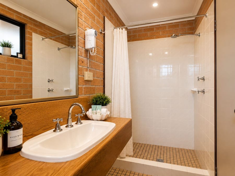 Un baño con tocador de madera, un lavabo blanco, un espejo, un secador de pelo montado en la pared y una zona de ducha con piso de baldosas.