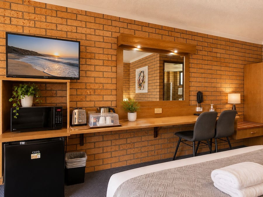 Habitación de hotel con pared de ladrillo, televisor, microondas, tostadora, refrigerador pequeño, escritorio con sillas y cama con toallas.