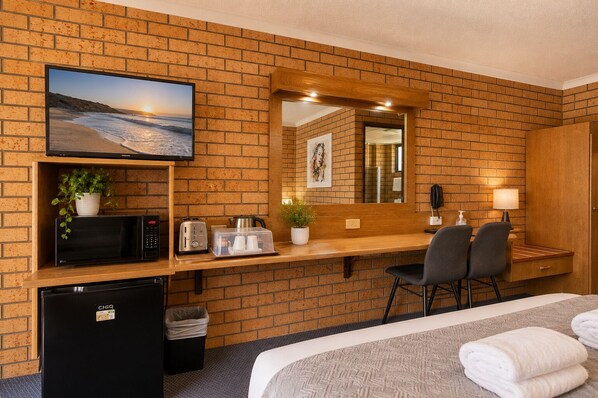 A hotel room with a brick wall, a television, a microwave, a toaster, a small refrigerator, a desk with chairs, and a bed with towels.