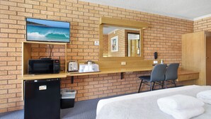 A hotel room with a brick wall, a TV, a microwave, a small fridge, a desk with a chair, and a bed. - Royal Palms Motor Inn (Coffs Harbour)