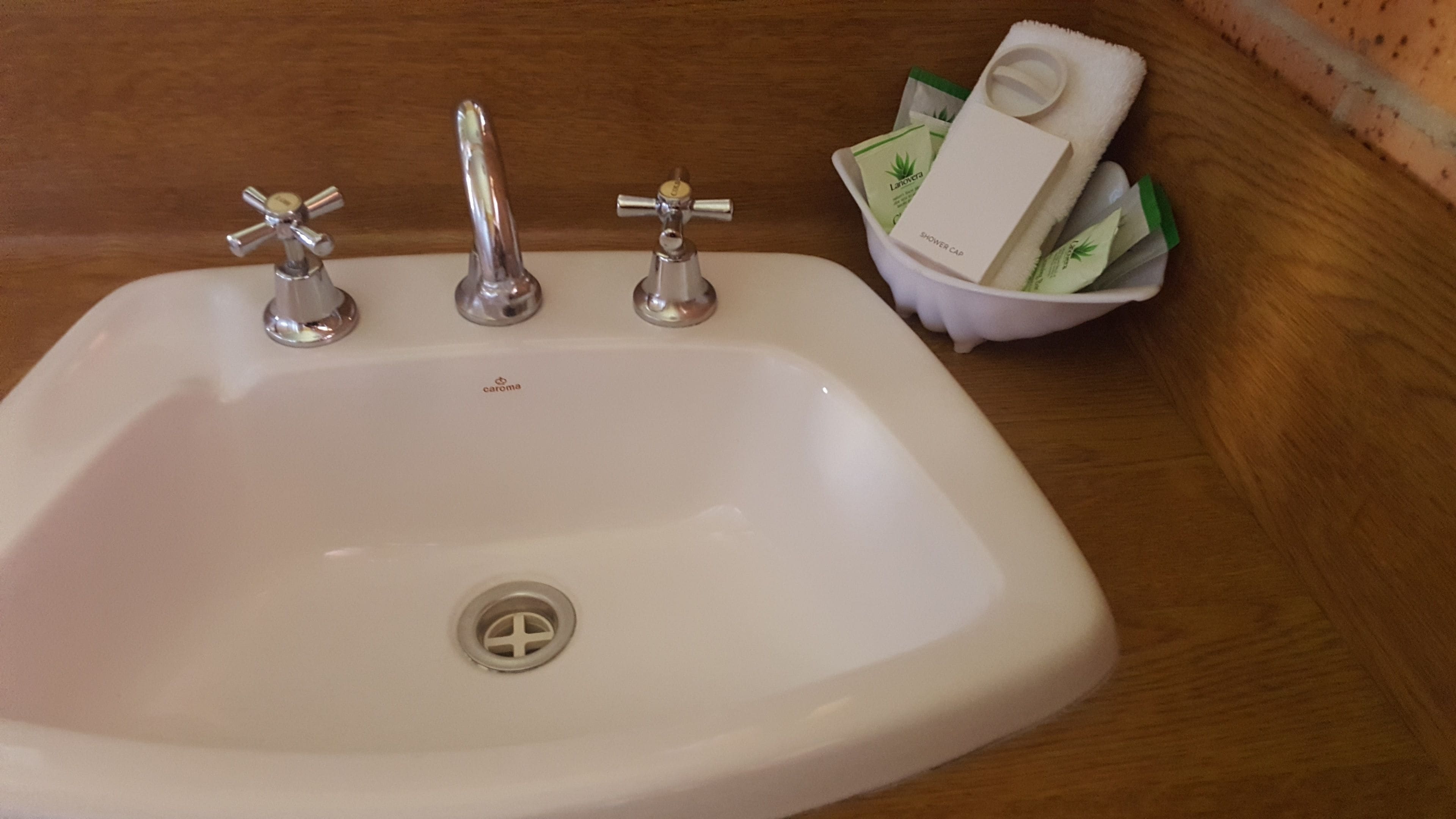 A white sink with three faucets, a drain, and a small basin containing a shower cap and toiletries.