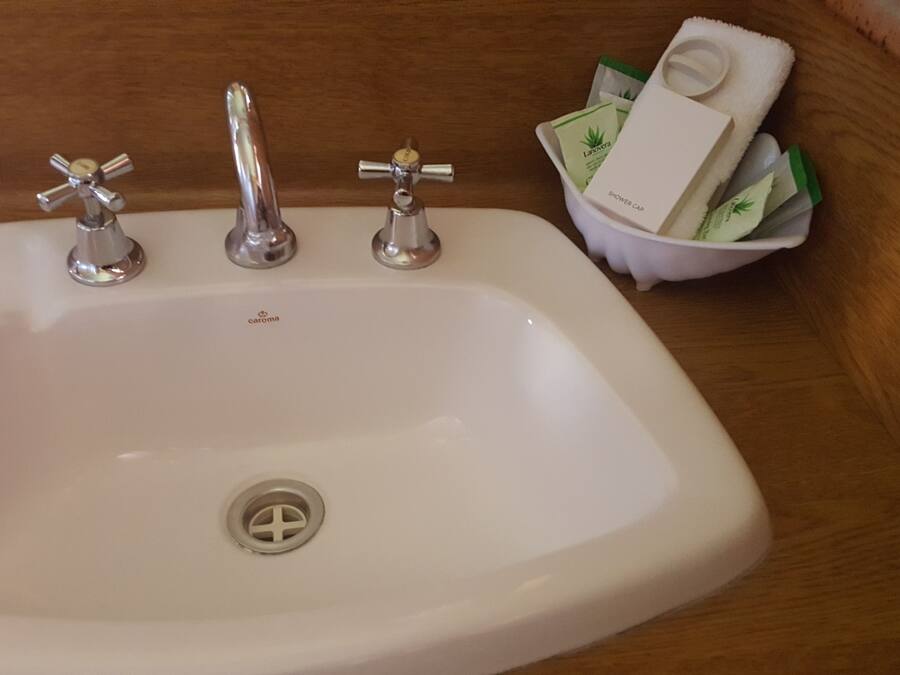 A white sink with three faucets, a drain, and a small basin containing a shower cap and toiletries.