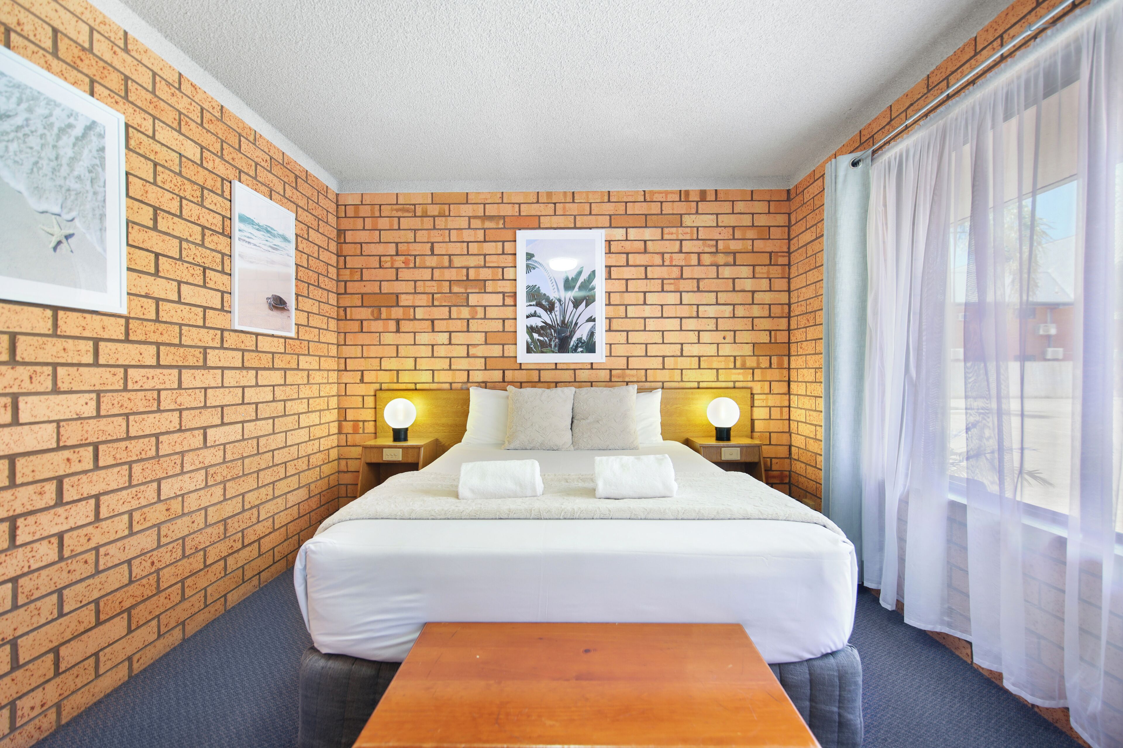 A hotel room with a brick wall, a bed with white bedding, two bedside lamps, and a window with curtains.