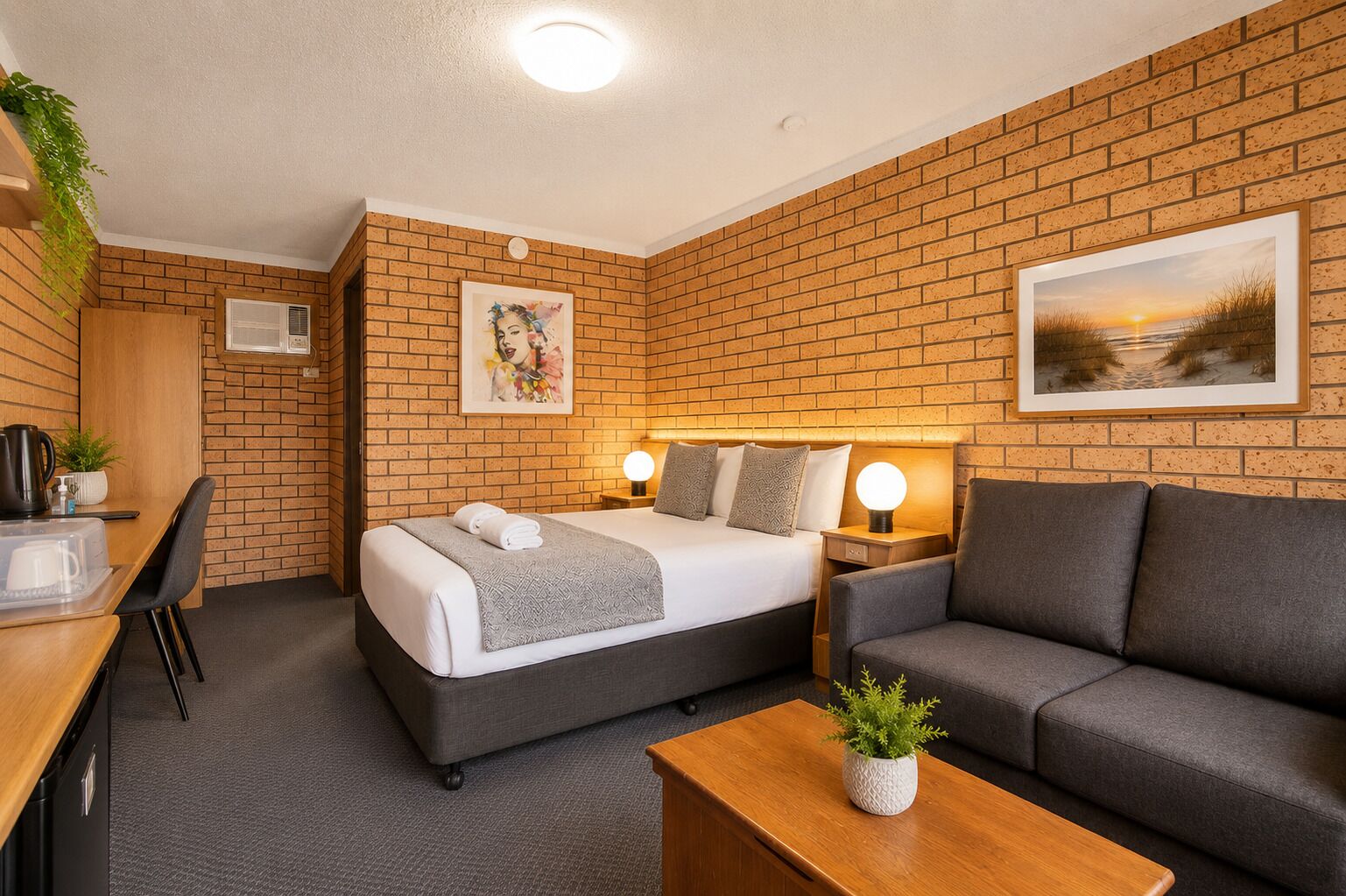 A hotel room with a brick wall, a bed with a grey headboard, a grey sofa, a wooden desk with a plant, and a picture of a sunset.