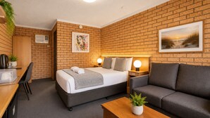 Habitación de hotel con pared de ladrillo, cama con cabecera gris, sofá gris, escritorio de madera con una planta y una imagen de un atardecer.