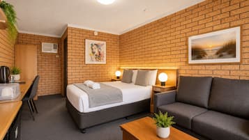 A hotel room with a brick wall, a bed with a grey headboard, a grey sofa, a wooden desk with a plant, and a picture of a sunset.
