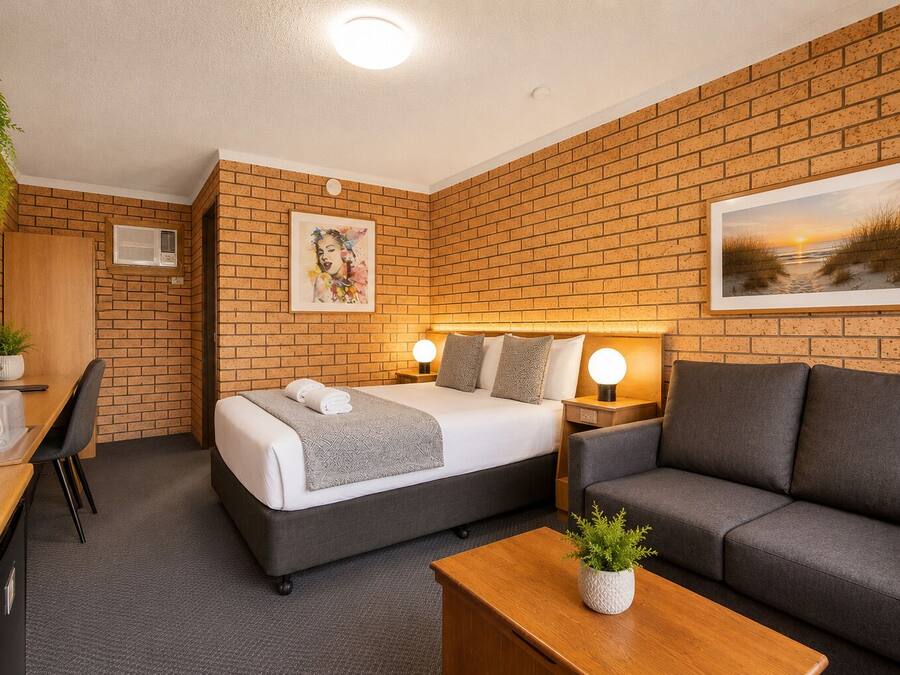A hotel room with a brick wall, a bed with a grey headboard, a grey sofa, a wooden desk with a plant, and a picture of a sunset.