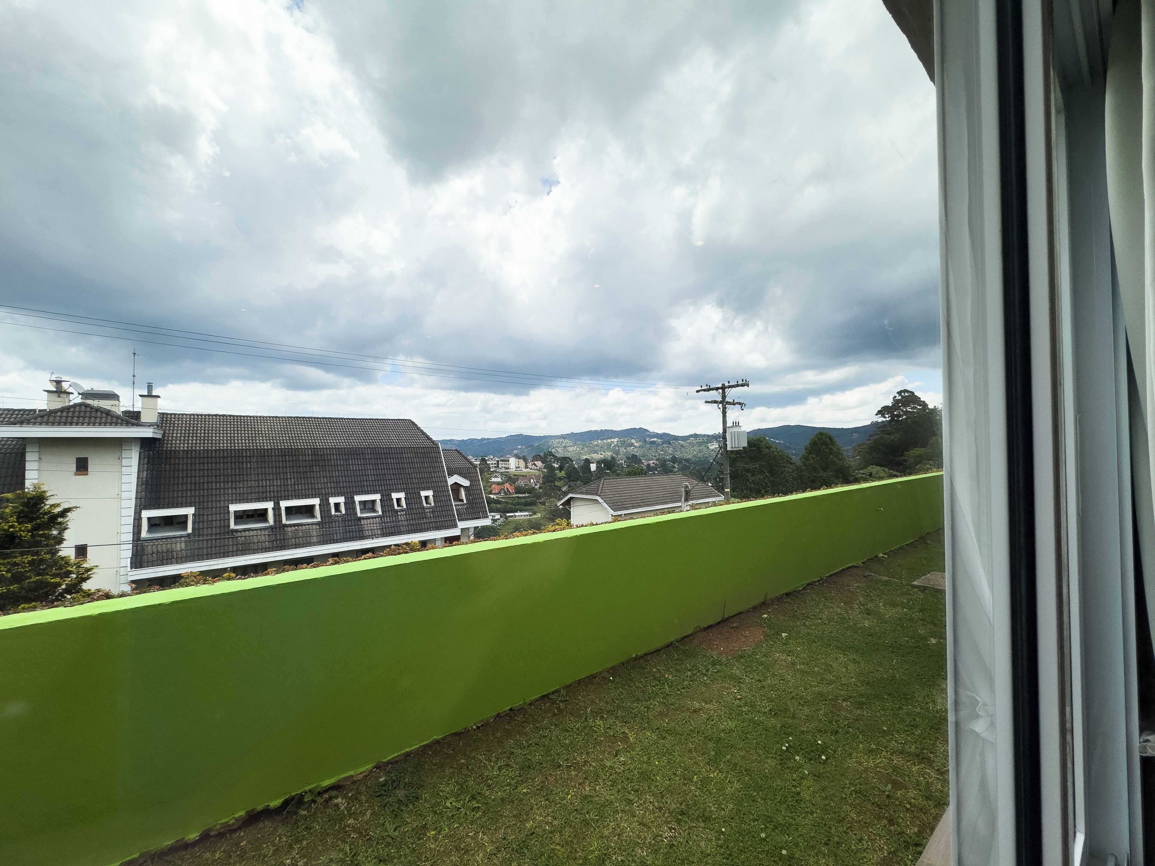 A view from a window overlooking a green barrier, a building with a dark roof, and a cloudy sky.