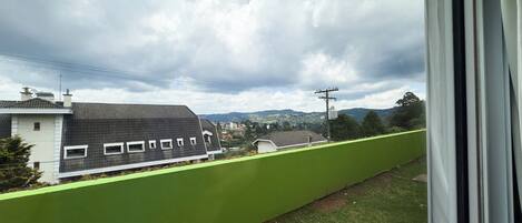 A view from a window overlooking a green barrier, a building with a dark roof, and a cloudy sky.