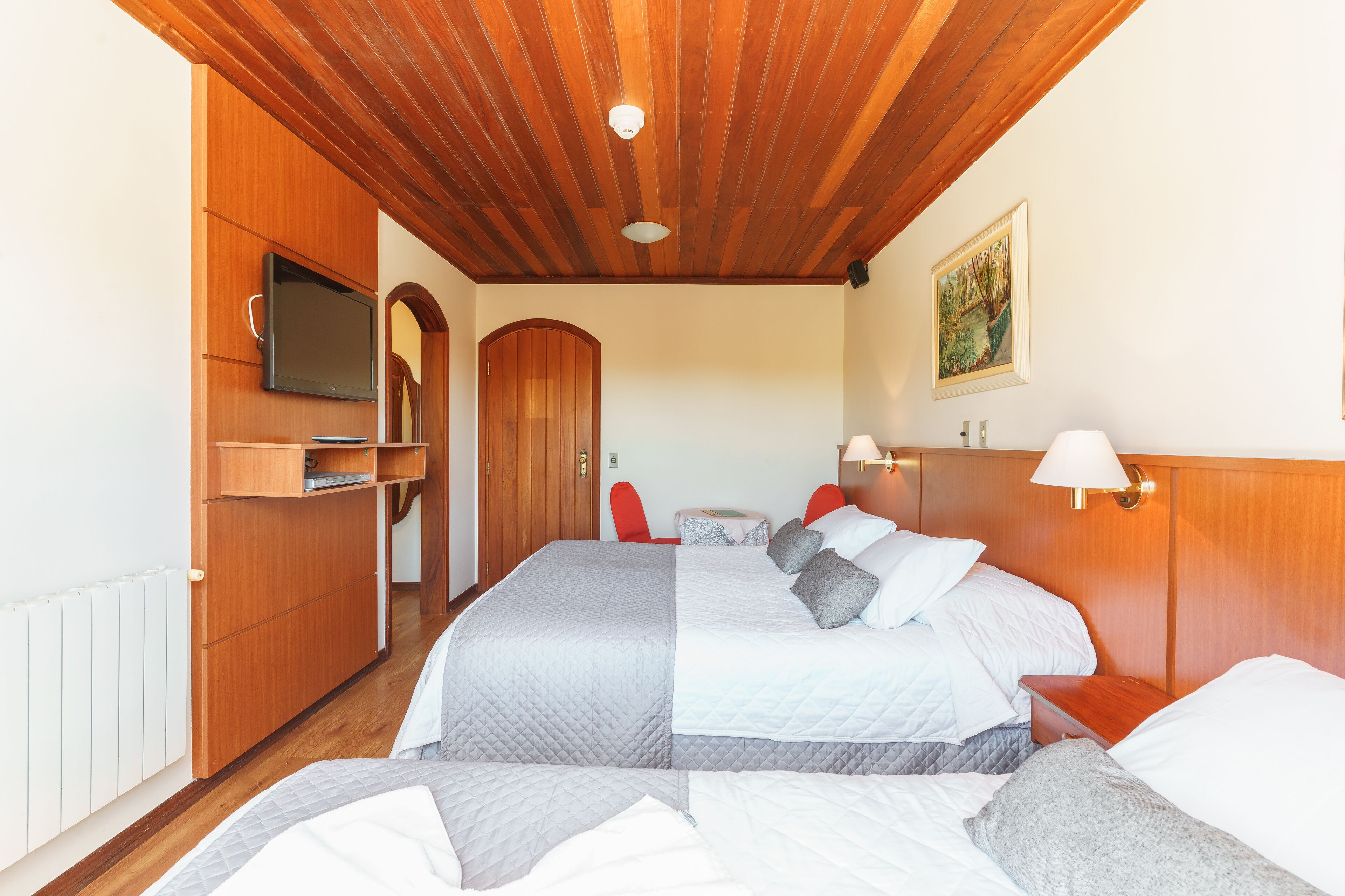 A hotel room with two beds, wooden ceiling, a TV, a painting, and a red chair.