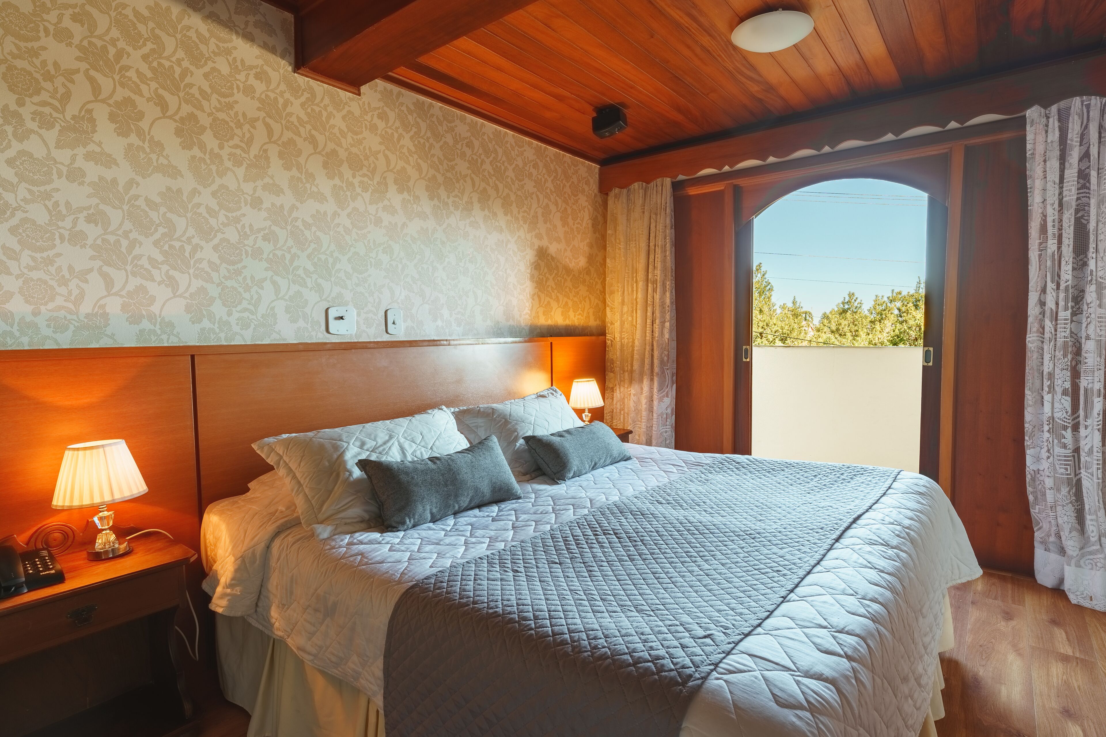 Une chambre d’hôtel avec un lit, deux lampes de chevet, un plafond en bois et une fenêtre avec des rideaux.