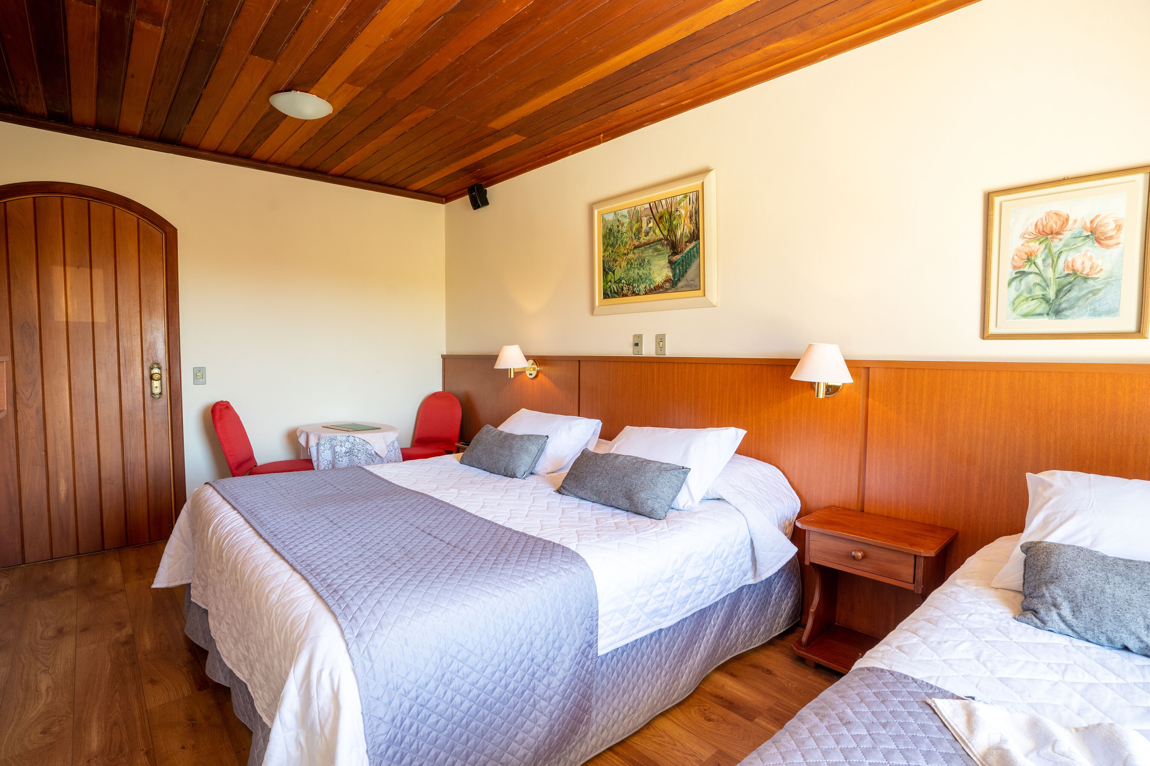 A hotel room with a wooden ceiling, a large bed with a quilted bedspread, two bedside tables with lamps, a red chair, and two framed floral paintings on the wall.