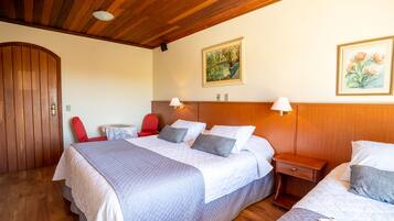 A hotel room with a wooden ceiling, a large bed with a quilted bedspread, two bedside tables with lamps, a red chair, and two framed floral paintings on the wall.