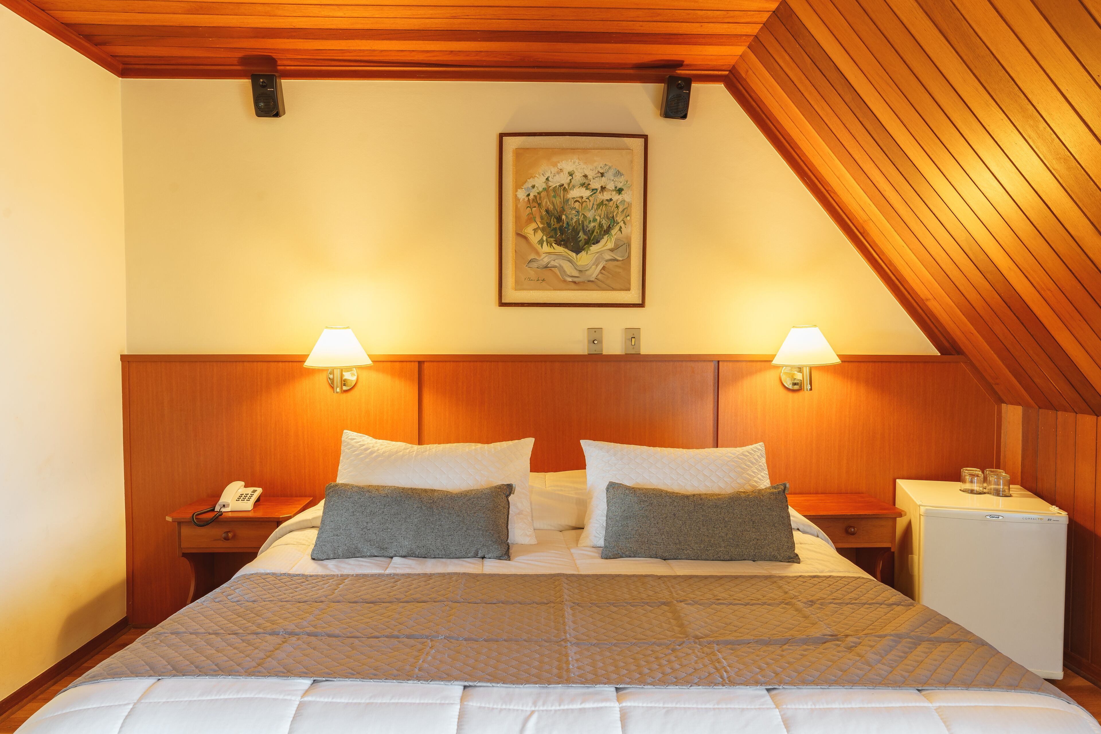 A hotel room with a large bed, two bedside lamps, a nightstand with a phone, a small refrigerator, and a framed artwork on the wall.