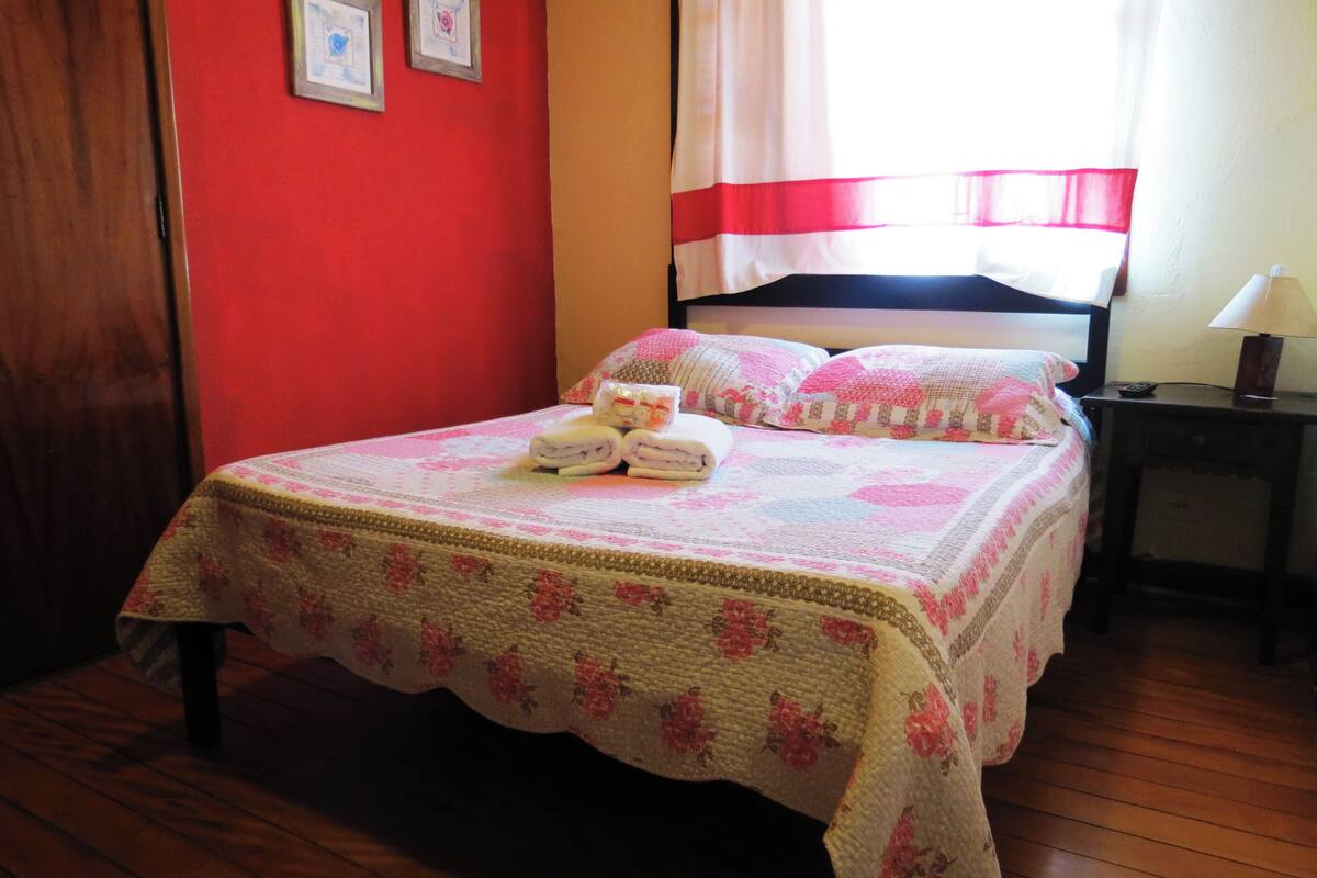 Una cama bien hecha con colcha estampada, una mesita de noche con lámpara y una ventana con cortinas.