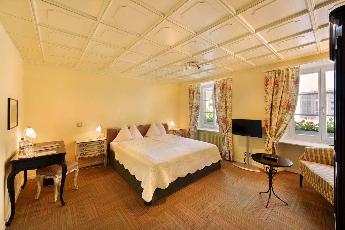 A hotel room with a large bed, a desk with a lamp, a chair, a small table, and a TV.