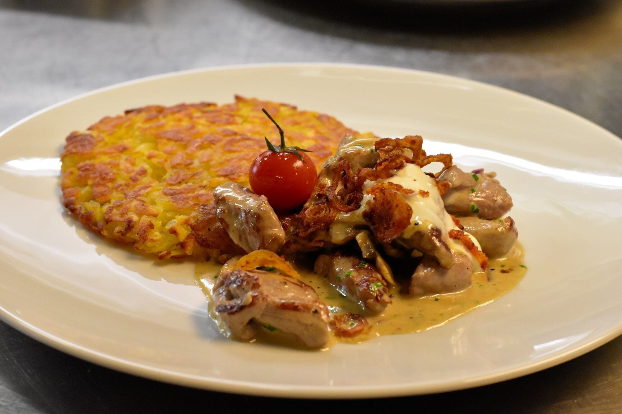 A plate with a dish consisting of a golden-brown pancake, a cherry tomato, and a creamy sauce with mushrooms.