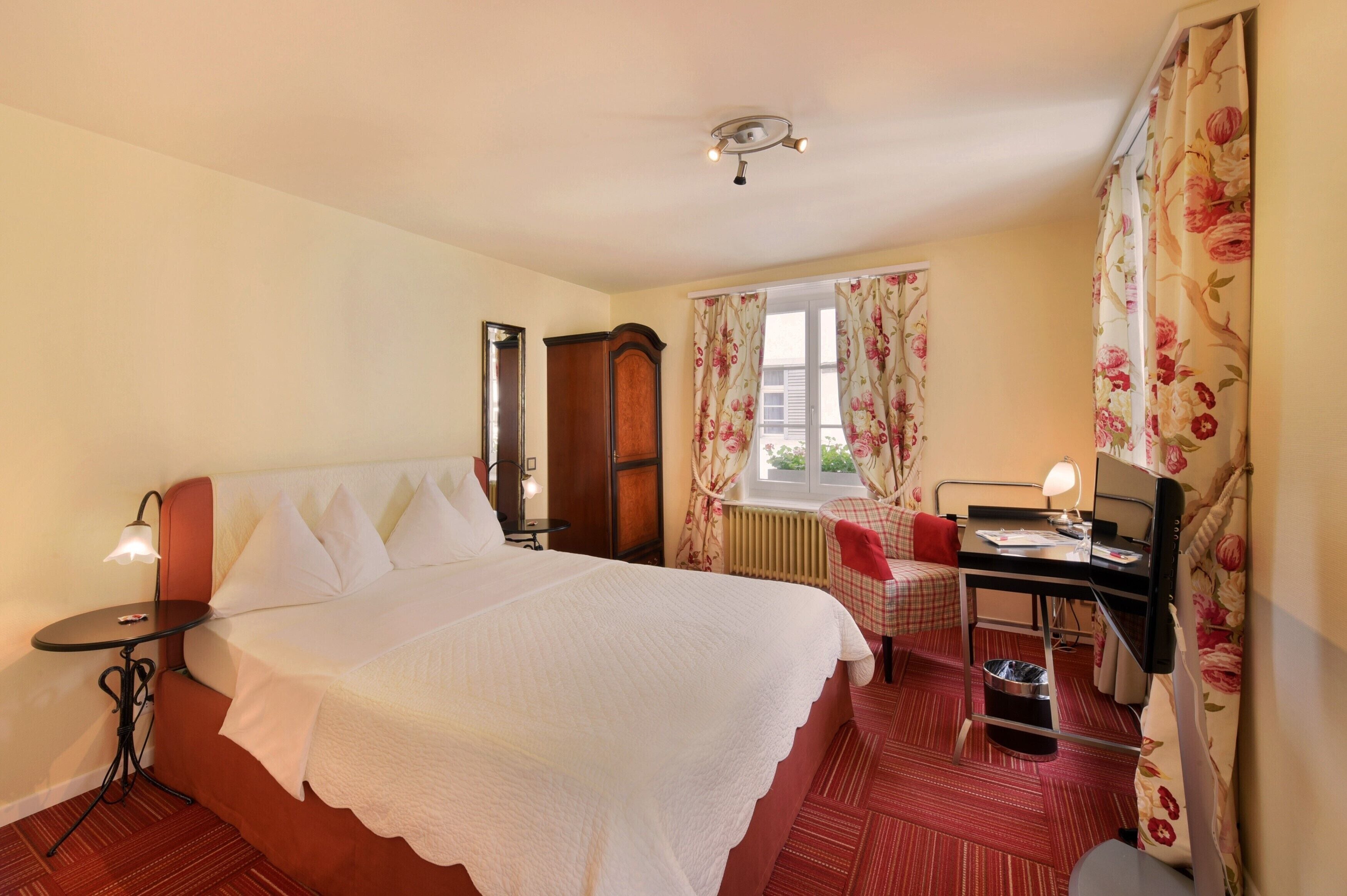 A hotel room with a large bed, a desk with a computer, a chair, a radiator, and a window with floral curtains.