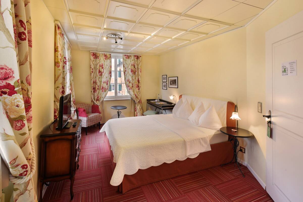 A hotel room with a bed, a desk, a chair, a small table, a lamp, and a window with curtains.