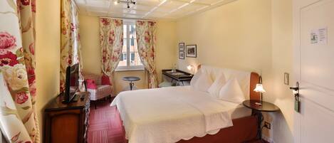 A hotel room with a bed, a desk, a chair, a small table, a lamp, and a window with curtains.