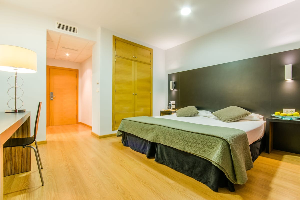 A modern hotel room with a bed, a desk, and a chair. There is a wardrobe and a nightstand with a lamp.