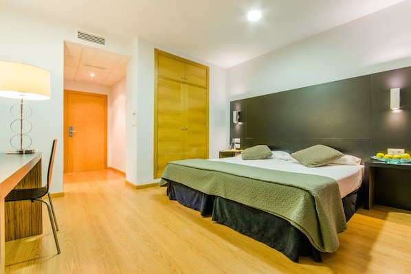 A modern hotel room with a bed, a desk, and a chair. There is a wardrobe and a nightstand with a lamp. - Hotel YIT Alcover (Granada)