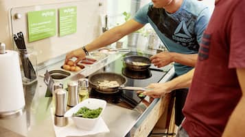 A kitchen counter with a frying pan, a bowl of green vegetables, and a container of eggs. There are two green signs on the wall, a metal container with utensils, and a white paper towel dispenser.
