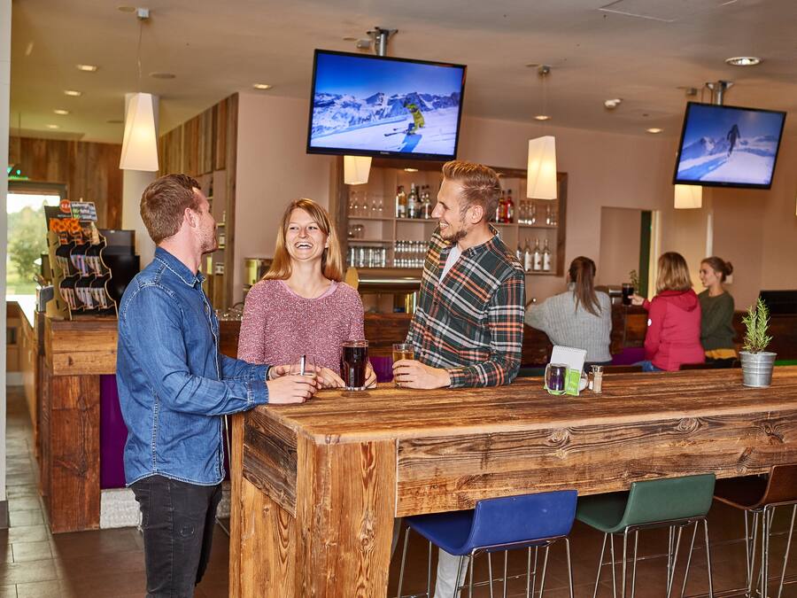A bar area with wooden bar counter, multiple screens displaying content, and seating with chairs.