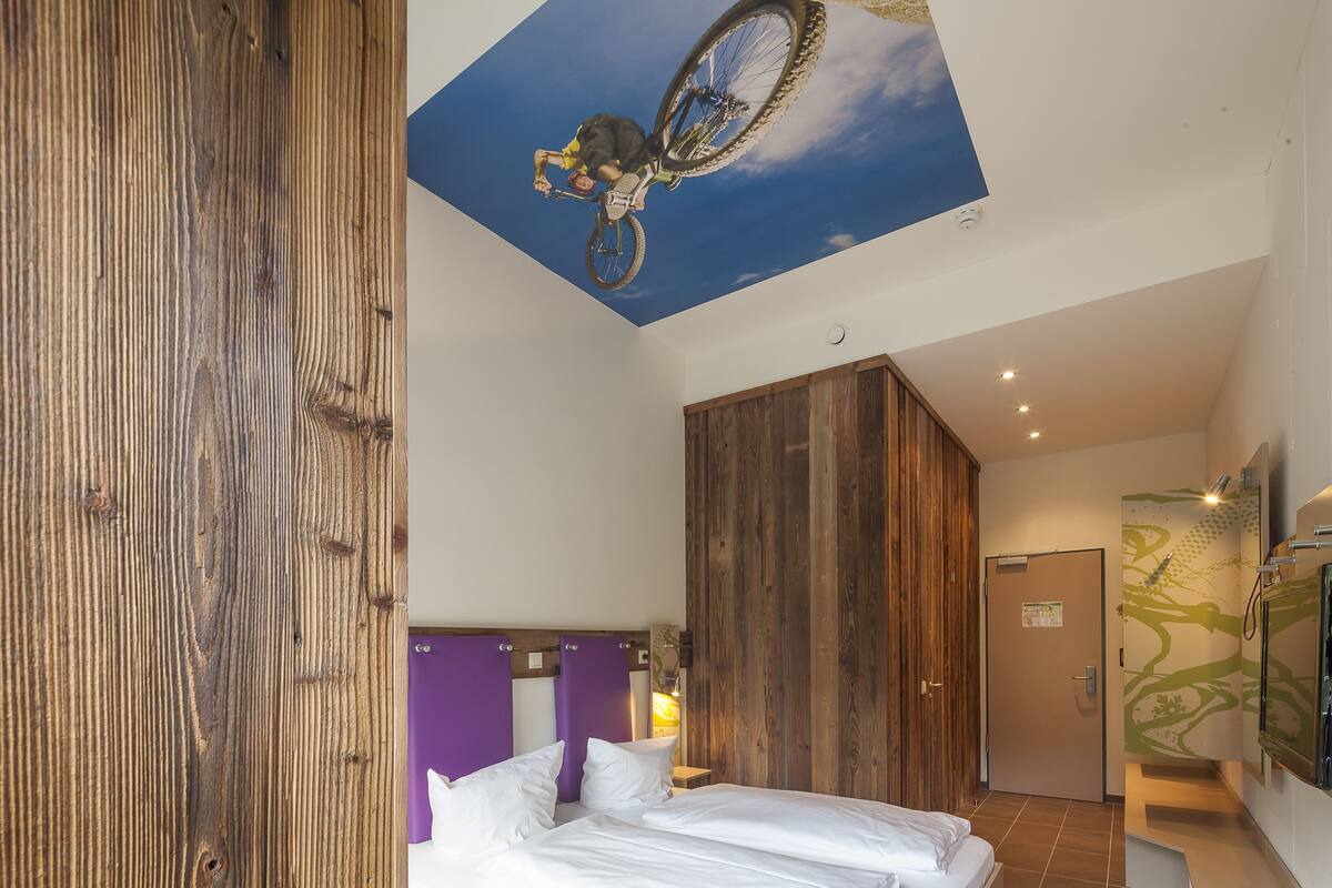 A modern hotel room with a large bed, wooden panel walls, a ceiling mural of a cyclist, and a door with a green tree design.