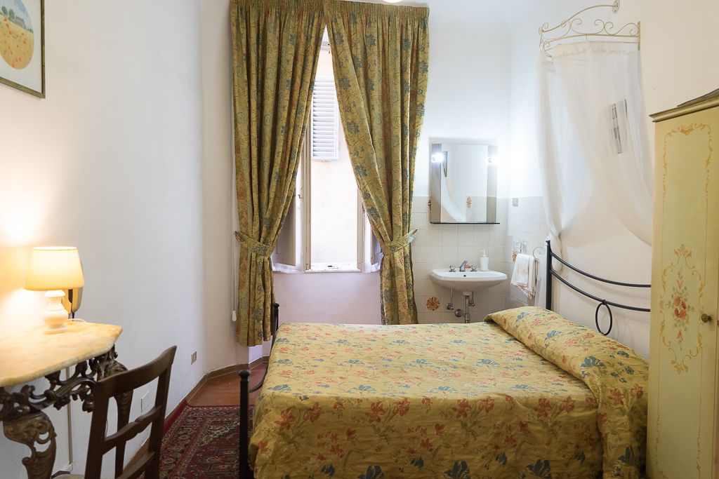 Un dormitorio con cama, mesita de noche con lámpara, ventana con cortinas, lavabo y espejo.