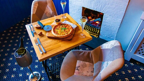 Un comedor acogedor con una pizza sobre una mesa de madera, una botella de vino y una chimenea al fondo.