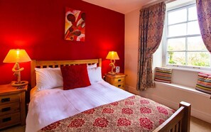 A hotel room with a red wall, a bed with white linens and a red pillow, two bedside lamps, a window with patterned curtains, and a floral-patterned throw on the bed.