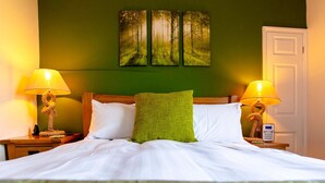 Una cama bien hecha con sábanas blancas, dos lámparas de noche, un cojín verde y un cuadro de un bosque en la pared.
