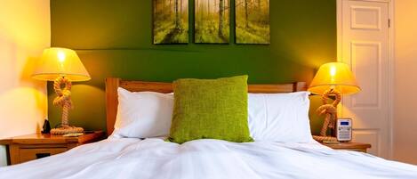 A neatly made bed with white linens, two bedside lamps, a green pillow, and a painting of a forest on the wall.